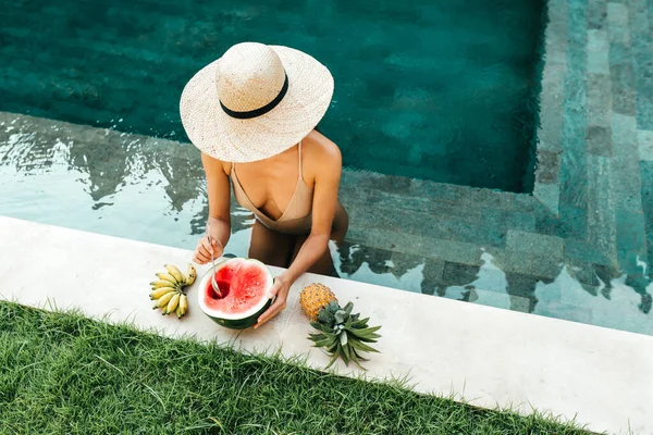 Jovem mulher atraente na piscina desfrutando de deliciosas frutas tropicais maduras: melancia, bananas e abacaxi. — Fotografia de Stock