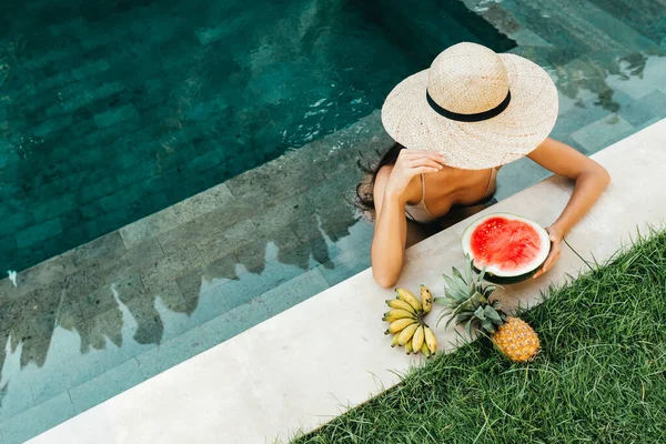Jovem mulher atraente na piscina desfrutando de deliciosas frutas tropicais maduras: melancia, bananas e abacaxi. — Fotografia de Stock