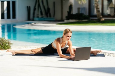 Genç, formda bir kadın laptopun önünde havuz kenarında yoga germe egzersizi yapıyor..