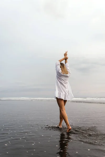 Mujer joven vistiendo hermoso vestido blanco está caminando en la playa durante la puesta del sol —  Fotos de Stock