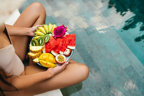 Ung oigenkännlig kvinna vid poolen med en tallrik tropiska frukter: vattenmelon, ananas, bananer, mangostan, passionsfrukt, mango och drakfrukt. — Stockfoto