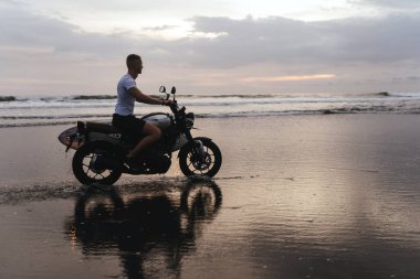 Gün batımında sörf tahtasıyla motosiklet süren sörfçü..