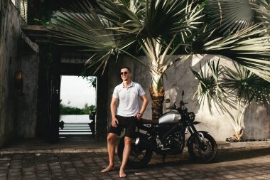 Güneş gözlüklü ve beyaz tişörtlü genç adam sokakta motosikletin yanında poz veriyor..