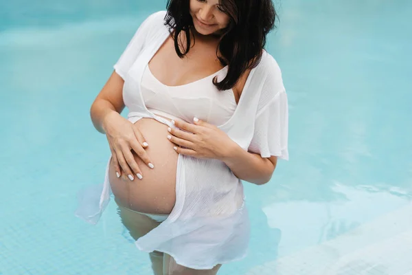 Zwangere vrouw ontspannen bij het zwembad en aanraken van haar buik met handen Stockfoto