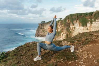 Uçurumun kenarında yoga yapan güzel bir kadın. Okyanusun muhteşem manzarasının arkasında.
