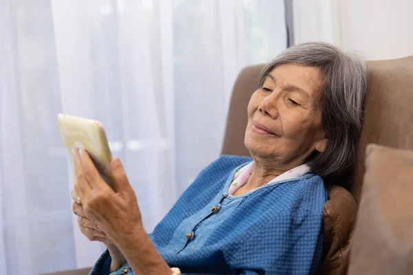 年长的亚洲女人在客厅的沙发上放松一下 在平板电脑上浏览无线互联网 — 图库照片