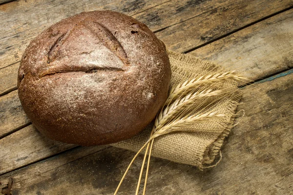 Loaf of rye bread with wheat ears on wood background — Stock Photo, Image