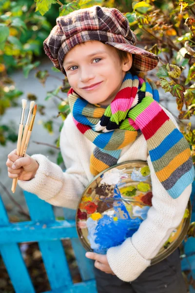Niño con pinturas en la paleta y pinceles cerca de la cerca —  Fotos de Stock