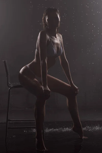 劇場の煙の中で黒のスタジオで水の雨の滴の下で椅子に座って白い下着の美しい濡れたブロンドの背の高い細い女の子 滑らかで健康的でしっとりとした肌 スペースのコピー — ストック写真