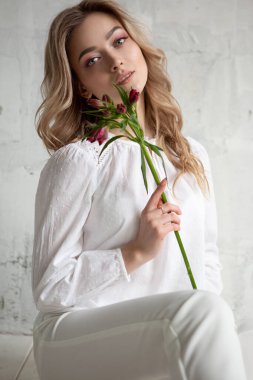 A young blonde woman with a flower in her hands is sitting on the floor. Fashion studio portrait