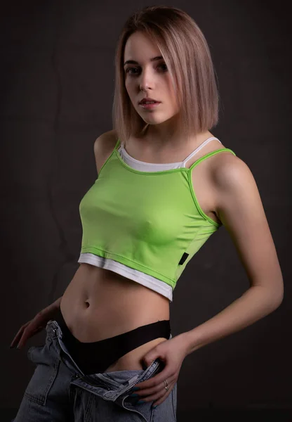 Gorgeous sexy blonde girl in a crop top and jeans poses on a dark background — Stock Photo, Image