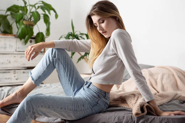 ファッショナブルなジャケットとジーンズにかわいい顔の美しい若いセクシーなブロンドがベッドの上に座っています ファッションスタジオの写真 — ストック写真