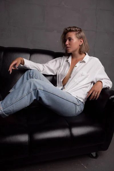 一个穿着白衬衫和牛仔裤的年轻貌美性感的金发姑娘在黑色沙发上摆姿势 黑暗背景下的工作室照片 — 图库照片