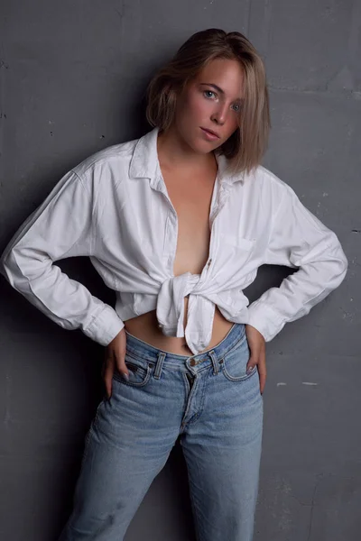 En vacker ung blond kvinna i en uppknäppt skjorta och jeans poserar på en grå bakgrund — Stockfoto
