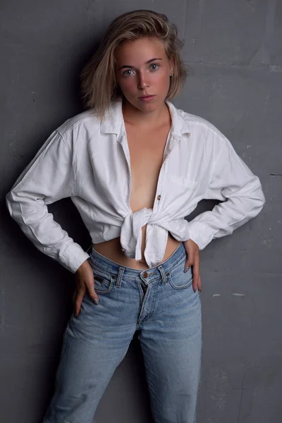 A gorgeous young blonde woman in an unbuttoned shirt and jeans poses on a gray background — Stock Photo, Image