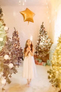Güzel küçük kız gülüyor ve elinde yıldız balonuyla Noel ağaçlarının yanında duruyor. Konsept mutlu çocukluk, yeni yıl ve doğum günü için dilek tutmak, dikey çekim