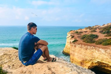Portekiz 'de taş üstünde gözlüklü, ince, yakışıklı bir adam oturuyor. Okyanusun arka görüntüsü