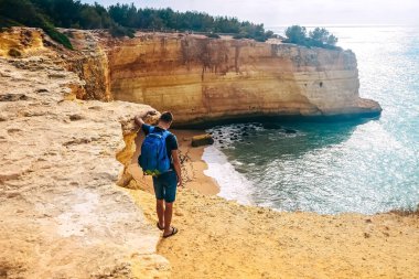 İnce bir adam Portekiz 'de bir kayanın üzerinde büyük bir sırt çantasıyla duruyor. Okyanusun arka görüntüsü