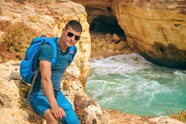 one slender guy sits with a big backpack on a rock in Portugal. guy looking at camera, ocean vie