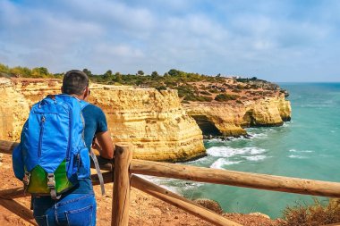 İnce bir adam Portekiz 'de bir kayanın üzerindeki çitin yanında büyük bir sırt çantasıyla duruyor. Arkadan okyanusa bakan bir adam mesafeye bakar.