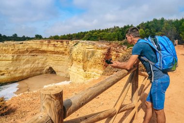 one thin guy stands with a large backpack near a fence on a cliff in Portugal with a camera in his hands. view from the back to the ocean, a man looks into the distanc