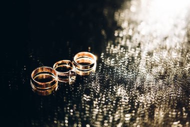 Ayna arka planında yan yana üç altın yüzük, siyah bir sırtüstü düğün aksesuarları.