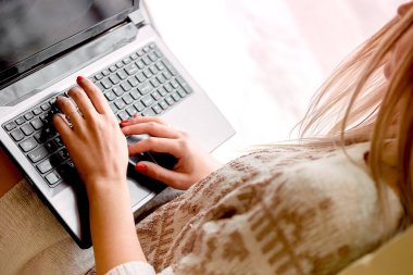 Hafif örülü kazaklı bir kız pencere eşiğinde oturuyor ve elinde dizüstü bilgisayar tutuyor.