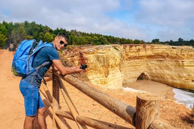 one thin guy stands with a large backpack near a fence on a cliff in Portugal with a camera in his hands. view from the back to the ocean, a man looks into the distanc