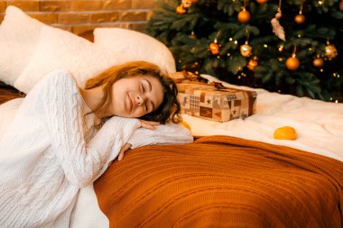 Siyah saçlı beyaz kazaklı güzel kıvırcık kız yatağa uzanıyor ve bir Noel ağacının arka planında uyuyor. Mutlu yıllar portresi, kız hayal kurar ve bir dilek tutar.