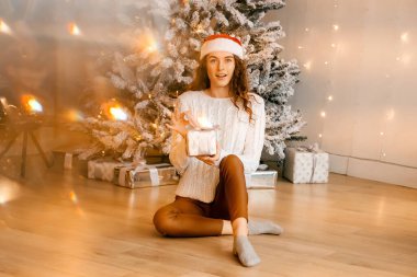 Siyah saçlı beyaz kazaklı güzel kıvırcık kız yere oturuyor ve elinde bir Noel ağacının arka planında bir hediye tutuyor. Mutlu yıllar, kızlar bir dilek diler.