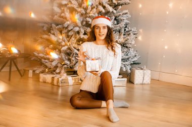 Siyah saçlı beyaz kazaklı güzel kıvırcık kız yere oturuyor ve elinde bir Noel ağacının arka planında bir hediye tutuyor. Mutlu yıllar, kızlar bir dilek diler.