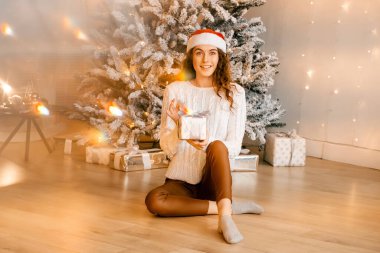 Siyah saçlı beyaz kazaklı güzel kıvırcık kız yere oturuyor ve elinde bir Noel ağacının arka planında bir hediye tutuyor. Mutlu yıllar, kızlar bir dilek diler.