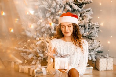 Siyah saçlı beyaz kazaklı güzel kıvırcık kız yere oturuyor ve elinde bir Noel ağacının arka planında bir hediye tutuyor. Mutlu yıllar, kızlar bir dilek diler.