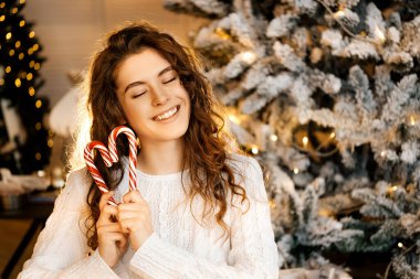 Kıvırcık saçlı güzel bir kız ağacın yanında duruyor, yeni yıl fotoğrafında şeker lolipopu var.