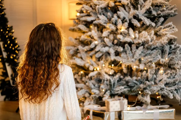 Siyah saçlı beyaz kazaklı güzel kıvırcık kız Noel ağacının yanında arkasını dönmüş oturuyor. Noel beklentisi, Yeni Yıl portresi