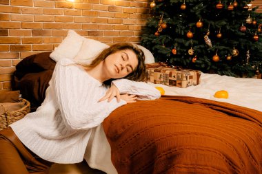 Siyah saçlı beyaz kazaklı güzel kıvırcık kız yatağa uzanıyor ve bir Noel ağacının arka planında uyuyor. Mutlu yıllar portresi, kız hayal kurar ve bir dilek tutar.