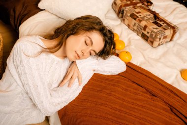 Siyah saçlı beyaz kazaklı güzel kıvırcık kız yatağa uzanıyor ve bir Noel ağacının arka planında uyuyor. Mutlu yıllar portresi, kız hayal kurar ve bir dilek tutar.