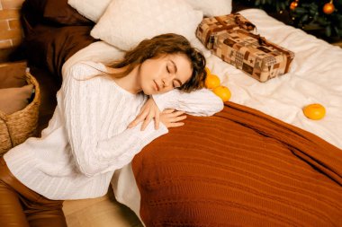 Siyah saçlı beyaz kazaklı güzel kıvırcık kız yatağa uzanıyor ve bir Noel ağacının arka planında uyuyor. Mutlu yıllar portresi, kız hayal kurar ve bir dilek tutar.