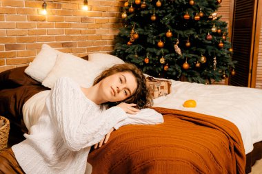 Siyah saçlı beyaz kazaklı güzel kıvırcık kız yatağa uzanıyor ve bir Noel ağacının arka planında uyuyor. Mutlu yıllar portresi, kız hayal kurar ve bir dilek tutar.