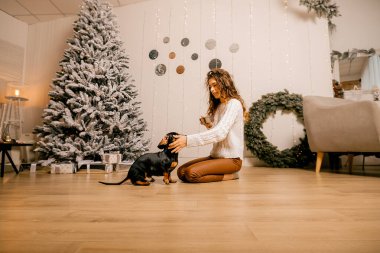 Beyaz kazaklı kıvırcık saçlı bir kız odada yerde oturuyor. Kadın ve köpek odada oynuyorlar. Kız köpeği besliyor..