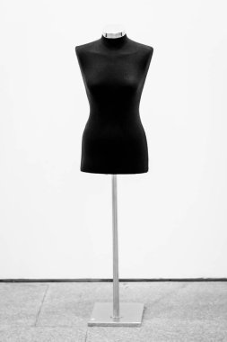 Kafası ve bacakları olmayan bir manken, siyah, arka planda beyaz bir duvar var..