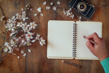 woman writing on blank notebook next to spring white cherry blossoms tree on vintage wooden table.