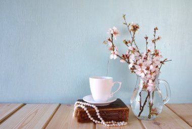 old book, pearls necklace and cup of coffee next to spring white flowers on wooden texture