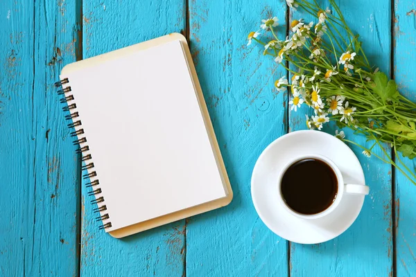 Bild von oben: Gänseblümchen-Blumen, leeres Notizbuch neben Tasse Kaffee auf blauem Holztisch. — Stockfoto