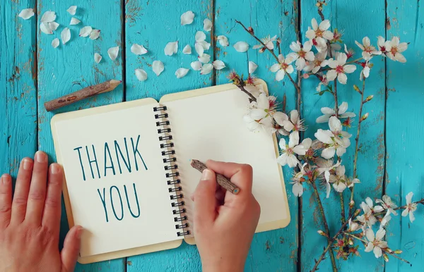 Vrouw hand het schrijven van een nota met de tekst dank u op een notebook, over houten tafel en kersenbloesem bloemen — Stockfoto