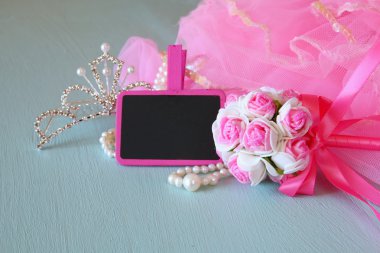 Small girls party outfit: crown and wand flowers next to small empty chalkboard on wooden table. bridesmaid or fairy costume