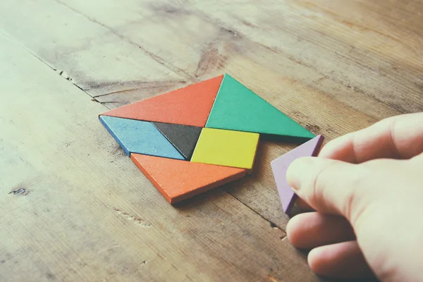 Mano del hombre sosteniendo una pieza perdida en un rompecabezas cuadrado tangram, sobre mesa de madera . — Foto de Stock