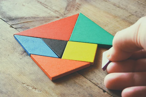 Man's hand holding a missing piece in a square tangram puzzle, over wooden table. — Stock Photo, Image