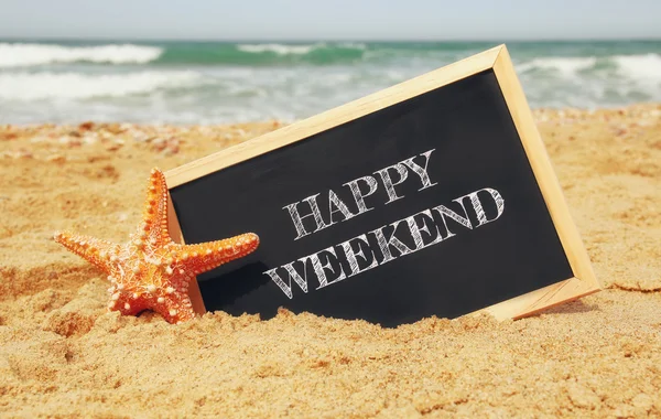 Starfish and clachboard with phrase: HAPPY WEEKEND, on sea sand and ocean horizon. selective focus — Zdjęcie stockowe