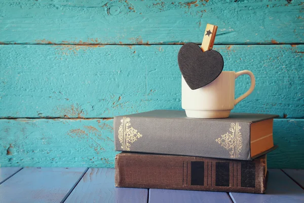 Coffee cup with little hart shape blank chalkboard and stack of old books on wooden table. vintage filtered — Stock Photo, Image